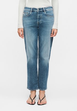 Kvinde iført lyseblå jeans med lige ben, hvid langærmet top og sorte thongsandaler, stående mod en ensfarvet baggrund.