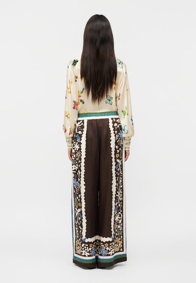 Γυναίκα με μακριά σκούρα μαλλιά στέκεται με την πλάτη γυρισμένη, φοράει μια κρεμ μπλούζα με λουλούδια και καφέ φαρδιά παντελόνια με πολύχρωμα διακοσμητικά μοτίβα στο τελείωμα.