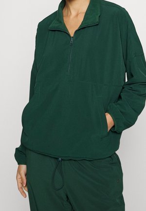 Person trägt dunkelgrünen langärmeligen Pullover mit halbem Reißverschluss und passende Hose, Hand in Pullovertasche, vor einfarbigem Hintergrund.