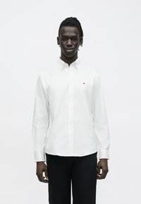 Jeune homme portant une chemise blanche à boutons avec un petit logo sur la poitrine et un pantalon noir, debout devant un fond blanc uni.