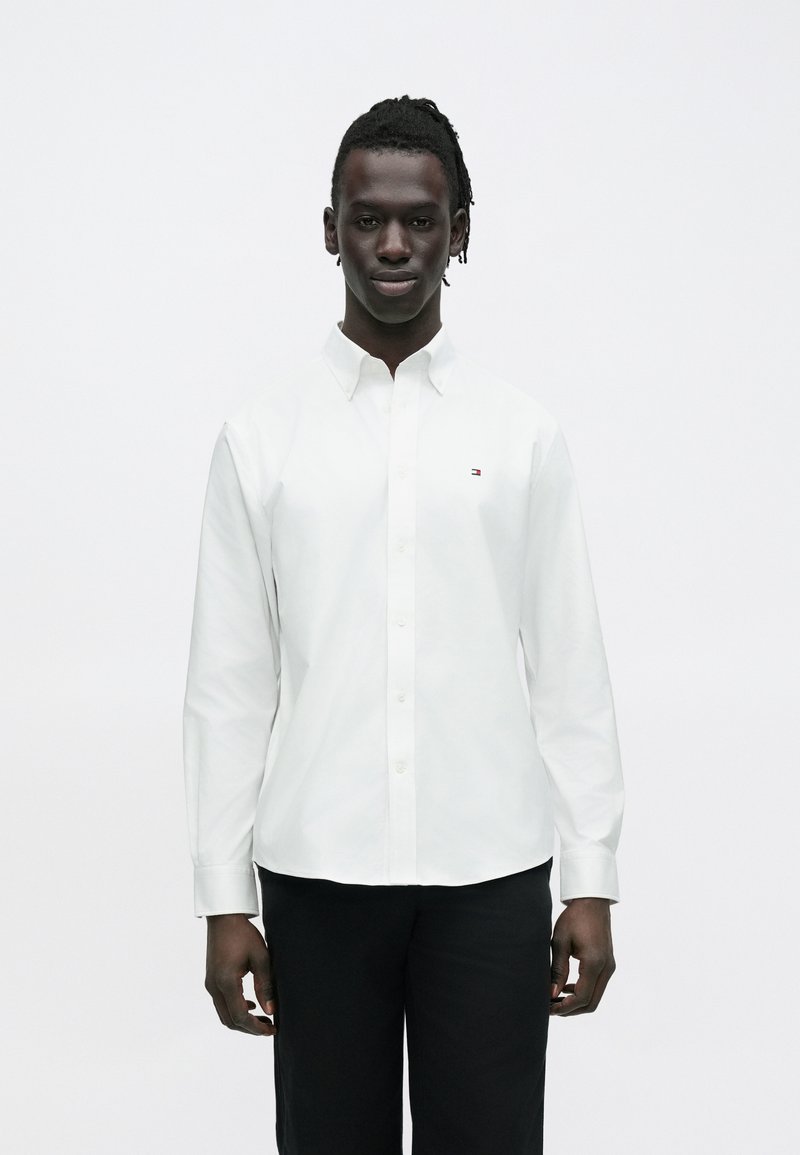 Jeune homme portant une chemise blanche à boutons avec un petit logo sur la poitrine et un pantalon noir, debout devant un fond blanc uni.