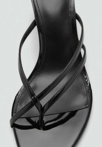 Sandalia de charol negro con un diseño sencillo. Presenta tiras entrecruzadas y una suela plana. Textura suave y acabado brillante.