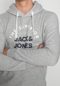 Sudadera gris con un bolsillo frontal, cordones blancos y un logotipo impreso. Tiene mangas largas y una textura suave.