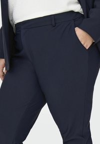 Tumši zili bikses, kas izgatavotas no gludas, nedaudz elastīgas auduma, ar sānu kabatām un tīrām šuvēm, nodrošinot piemērotu piegriezumu.