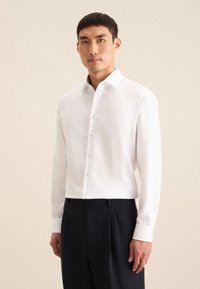 Camicia bianca a maniche lunghe con colletto strutturato, patta dei bottoni frontale e polsini in contrasto, abbinata a pantaloni formali scuri.