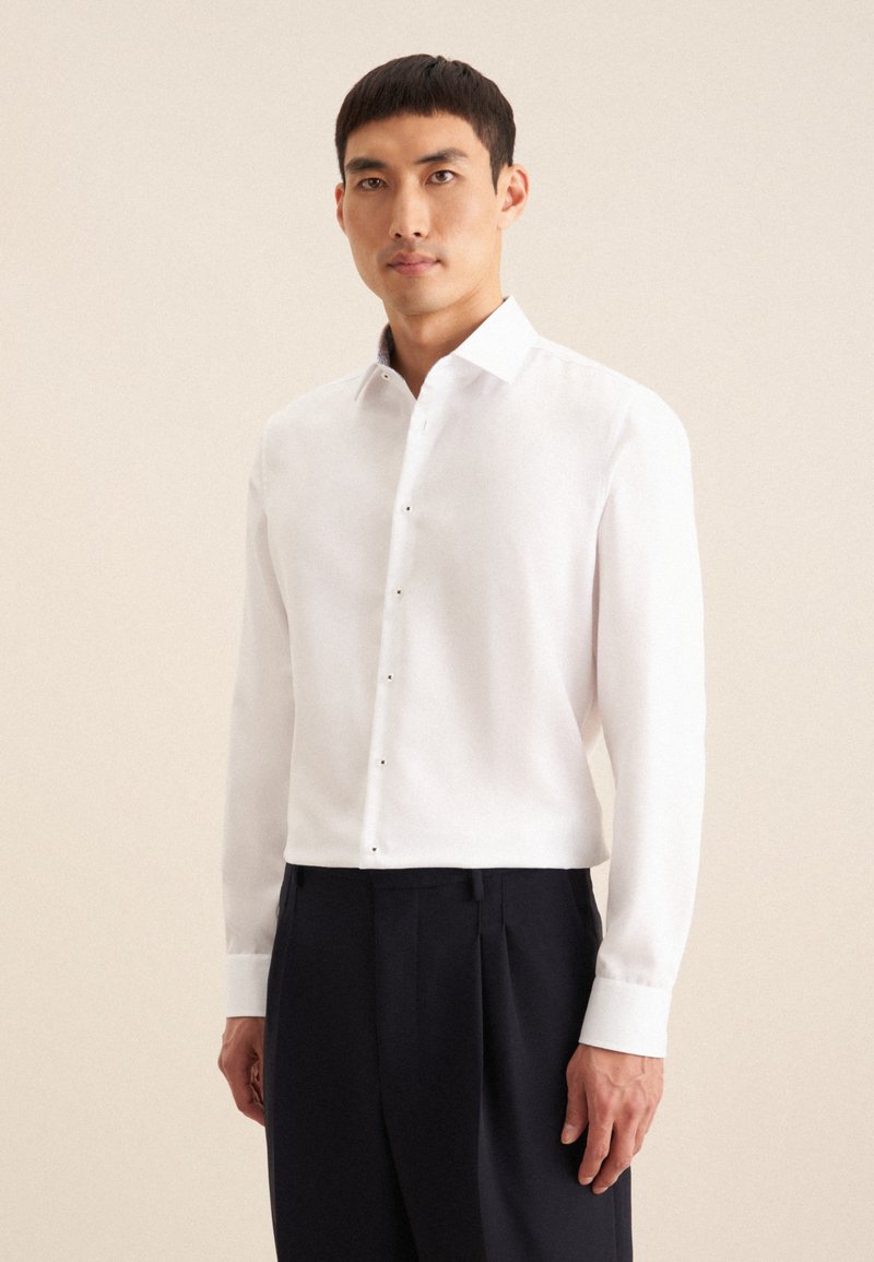 Camicia bianca a maniche lunghe con colletto strutturato, patta dei bottoni frontale e polsini in contrasto, abbinata a pantaloni formali scuri.
