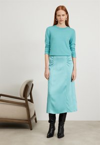 Pull en tricot turquoise à manches longues associé à une jupe midi en satin assortie avec ruchés sur les côtés, porté avec des bottes noires hauteur genoux.