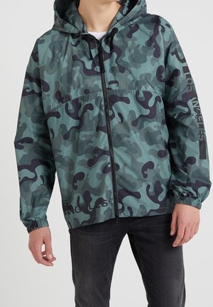 Persona che indossa una giacca con cappuccio verde e nera mimetica con cerniera e polsini elastici, e pantaloni neri.