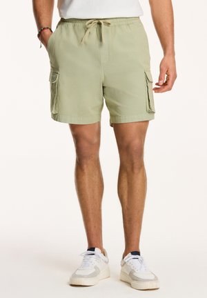 Shorts cargo vert clair avec taille élastique et cordon de serrage, dotés de poches latérales et d'un tissu texturé. Portés avec des baskets blanches.
