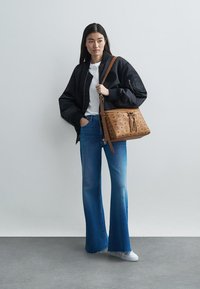 Barna táska perforált mintázattal, csatolt pánttal, fekete bomber dzseki, fehér póló, kék trapéz farmer és fehér sportcipő.