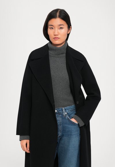 Femme portant un long manteau noir sur un pull à col roulé gris et un jean bleu, avec une main dans la poche, se tenant contre un fond uni.