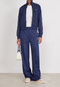 Chándal azul marino con rayas laterales blancas, chaqueta con cremallera, pantalones de corte relajado y un bolso blanco, combinado con zapatillas beige.