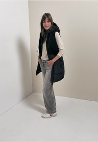 Schwarze gepolsterte Weste mit Kapuze, über einem hellen Pullover getragen, kombiniert mit hellgrauen Jeans und weißen Sneakers mit roten Akzenten.