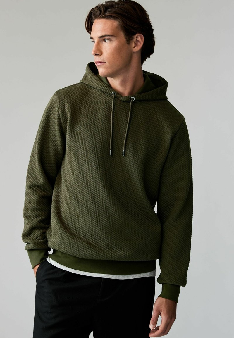 Olijfgroene hoodie van een gestructureerde, textuurstof, met een trekkoord capuchon en geribde manchetten. Gelaagd over een wit shirt.