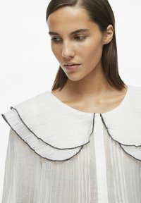 Blusa blanca con un cuello de volantes de doble capa con borde negro, textura de pliegues verticales y un diseño de corte recto.