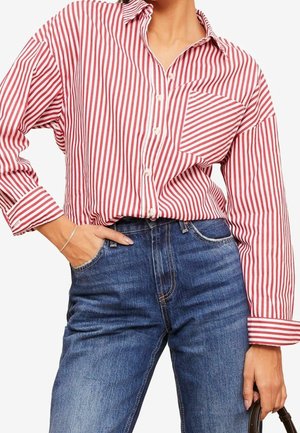 Kobieta ubrana w czerwono-białą koszulę z pionowymi paskami, zapinaną na guziki, z kieszenią na piersi, włożoną do wysokich niebieskich dżinsów, z ręką w kieszeni.