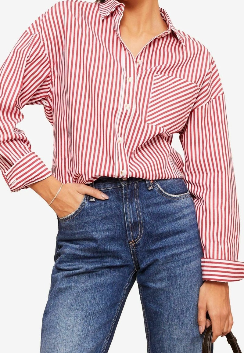 Nainen, joka on pukeutunut puna-valkoisiin pystyraitoihin olevaan napilliseen paitaan, jossa on rintatasku, ja jonka hän on pistänyt korkeavyötäröisiin sinisiin farkkuihin, käsi taskussa.