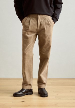 Beige corduroy trousers with pleats, two side pockets, and a button closure, paired with dark brown ankle boots on a wooden floor.