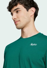Camiseta verde de cuello redondo hecha de algodón con una textura suave; presenta un pequeño logotipo blanco de Adidas en el área del pecho izquierdo.