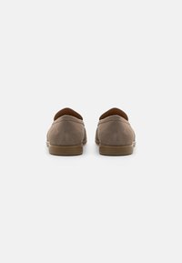 Chaussures à enfiler en suède marron avec un bout arrondi, un col rembourré et une semelle en caoutchouc beige, présentant une texture lisse et des détails de couture subtils.
