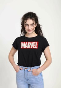 Camiseta de algodón negra con mangas cortas enrolladas, que presenta un gran logo "MARVEL" en rojo y blanco impreso en el frontal. Combinada con jeans de un azul claro.