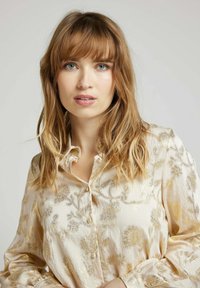 Blouse en tissu beige clair avec un motif floral en métal doré, fermeture à boutons et manches longues avec poignets froncés.