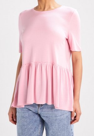 Femme portant un haut rose clair à manches courtes ample avec une taille froncée et un jean en denim bleu sur fond uni.