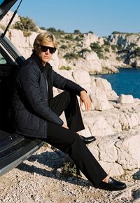 Homme portant des lunettes de soleil et une veste matelassée sombre assis au coffre ouvert d’une voiture sur des falaises côtières rocheuses au bord d’une eau bleue.