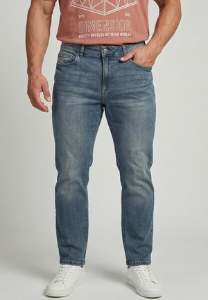 Mann trägt blaue ausgewaschene Jeans, weiße Turnschuhe und ein rotes T-Shirt mit geometrischem und Text-Design, steht mit einer Hand in der Tasche.