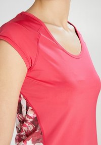 Primer plano de una persona que lleva una camisa de manga corta rosa con paneles laterales de estampado floral y un escote en forma de barco.