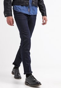 Tummansinisiä räätälöityjä housuja, joissa on vaaleansininen paita ja musta farkkuliivi, yhdistettynä mustiin brogue-kengiin. Siisti, moderni istuvuus.