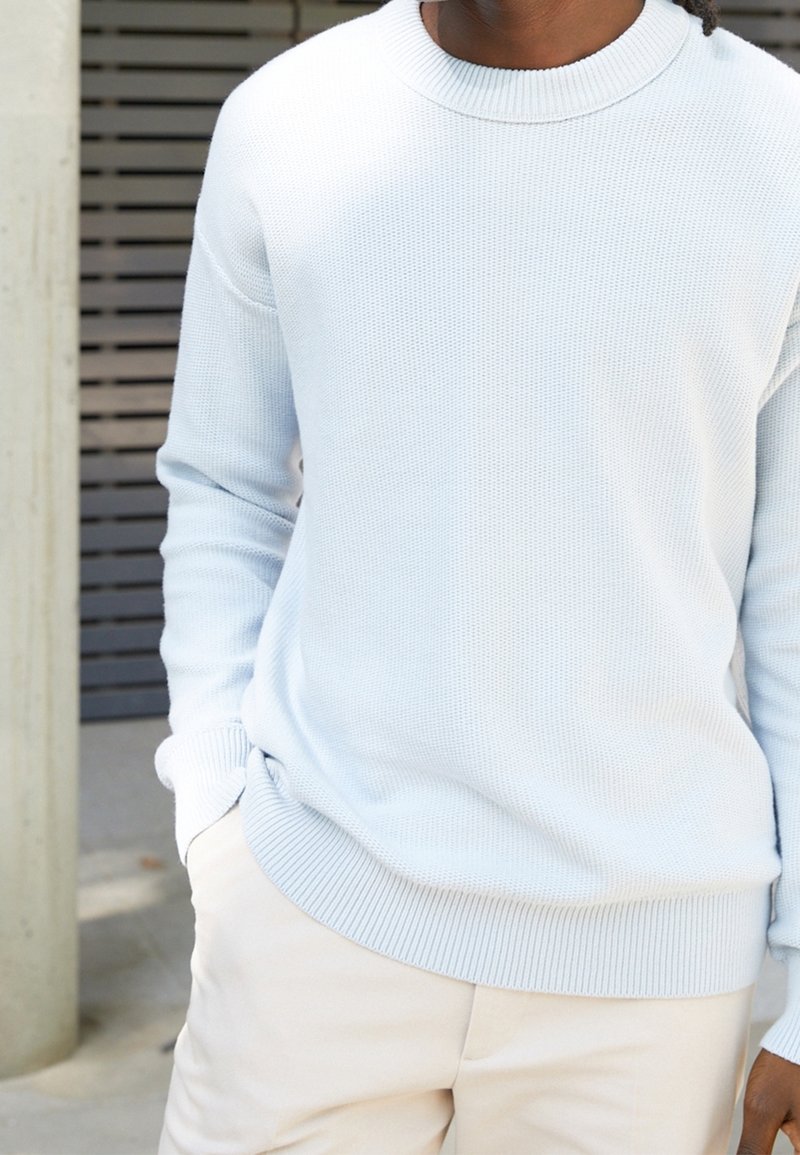 Hellblau gestrickter Rundhals-Pullover mit gerippten Bündchen und Saum, der ein strukturiertes Muster aufweist. Kombiniert mit hellen Hosen.