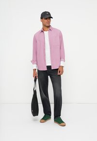 Helles rosafarbenes Hemd mit Knöpfen, weißes T-Shirt, graue Mütze, schwarze Jeans, grüne Sneakers und eine schwarze Tasche mit Logo dabei.