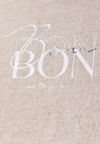Tejido beige con letras blancas en relieve que dicen "BON" y un acento en estilo manuscrito que dice "cosas buenas por venir" en texto más pequeño. Textura suave.
