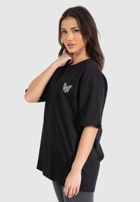 Sort, oversized t-shirt i bomuld med korte ærmer, der har et sølvfarvet sommerfuglemotiv på venstre bryst. Afslappet pasform og glat struktur.