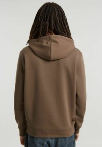Felpa con cappuccio marrone dal tessuto morbido e con polsini a costine. Presenta un'etichetta cucita sul lato sinistro in basso, abbinata a jeans blu.