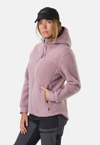 Woman wearing a light purple fleece jacket with hood and zip, black cap, and dark gray pants with zip pockets, posing with one hand in jacket pocket.