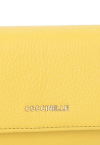 Portafoglio giallo in pelle con texture fine, con logo "Coccinelle" in argento e cuciture ordinate lungo i bordi. Forma rettangolare compatta.