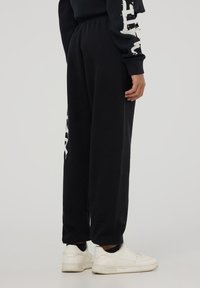 Czarne dresy z elastycznym pasem, luźny krój, z wyraźnym białym napisem i grafikami na lewej nogawce i boku.