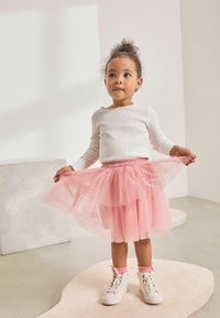 Ein Kind trägt ein weißes Langarmshirt und einen pinken Tüllrock. Kombiniert mit weißen Sneakers und pinken Socken. Heller Hintergrund.