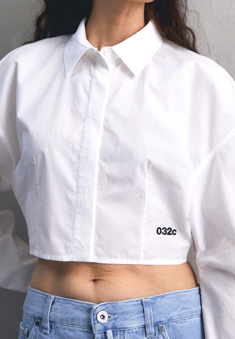 Afgeknot wit shirt met een button-down kraag, gemaakt van gladde stof, met een zwart logo "032c" rechts onderaan.