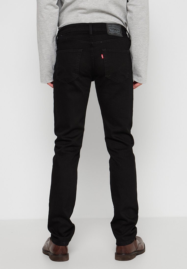 Persona vestida con jeans negros de Levi's, una camiseta de manga larga gris y zapatos de vestir marrones, de pie frente a un fondo blanco.