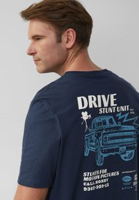 Marineblauwe T-shirt met een grafisch ontwerp op de achterkant, waarop een illustratie van een vrachtwagen, filmcamera en tekst in wit en lichtblauw is afgebeeld.