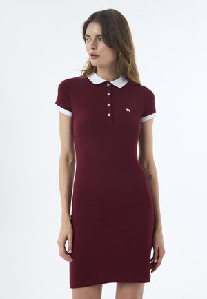 Femme portant une robe polo bordeaux ajustée avec col et poignets blancs, regardant vers sa gauche sur un fond uni.
