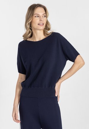 Blondynka w granatowym swetrze z krótkim rękawem i dopasowanych spodniach, stojąca z jedną ręką na biodrze, patrząca na bok na białym tle.