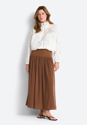 Naine kannab valget lukuga jopet, millel on lõigatud mustrid, kõrge vöökohaga pruuni maxi seelikut ja musti kontsaga sandaale, seistes valgel taustal.