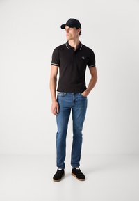 Fekete póló fehér kontrasztos díszítéssel, slim-fit kék farmernadrág és fekete cipő. Kiegészítve egy fekete baseball sapkával.