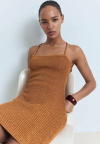 Abito a maglia marrone a coste con spalline sottili, silhouette aderente, che arriva a metà coscia. Accompagnato da un bracciale bordeaux al polso.