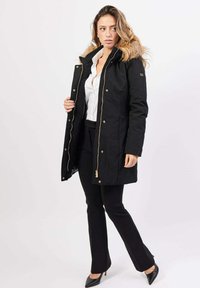 Donna con capelli lunghi che indossa un parka nero con cappuccio di pelliccia, una camicia bianca, pantaloni neri e tacchi neri, in piedi di fronte a uno sfondo chiaro e uniforme.