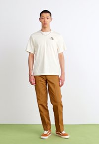 Lys beige t-skjorte med et lite grafisk trykk, kombinert med tan bukser og oransje sneakers med hvite detaljer. Minimalistisk design og passform.